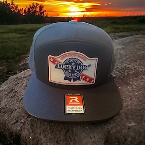 May include: A navy blue flat bill snapback hat with a mesh back. The hat features a patch with the text "LUCKY DOG GUITARS" and "HANDCRAFTED IN TENNESSEE". The hat also has a "RICHARDSON" tag.