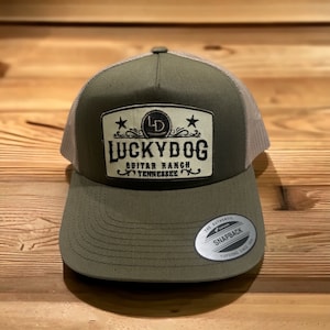 Könnte beinhalten: Eine grüne und graue Trucker-Kappe mit einem braunen Patch, auf dem "Lucky Dog Guitar Ranch Tennessee" steht, mit einem Stern auf jeder Seite des Patches. Der Patch hat auch einen goldenen Kreis mit den Buchstaben "LD" darin.