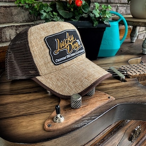May include: A brown and tan trucker hat with a mesh back and a patch that reads "Lucky Dogs Custom Guitars Cleveland." The hat is resting on a custom guitar with a wood grain finish.