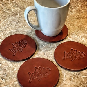 May include: A light blue mug rests on a brown leather coaster, with three additional coasters nearby. Each coaster is round and embossed with the words "Lucky Dogs" in a script font, along with a dog paw print.
