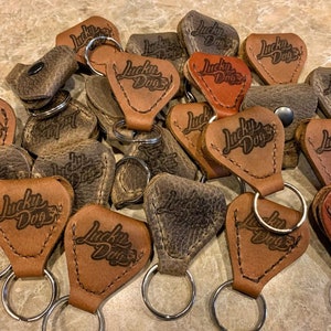 May include: A close-up of a pile of leather guitar picks with the words "Lucky Dogs" embossed on them. The picks are in various shades of brown and have a distressed look. Each pick has a silver key ring attached.