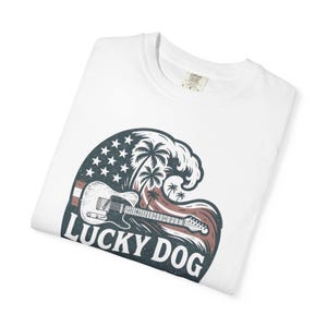 May include: White t-shirt with a graphic featuring a guitar, wave, and palm trees. The design incorporates the American flag and the text "LUCKY DOG" in a distressed font. The shirt is folded.