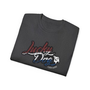 May include: A dark gray t-shirt with a red, white, and blue graphic that says "Lucky Dog" with a small white dog silhouette. The text "Custom Guitars" is below the graphic.