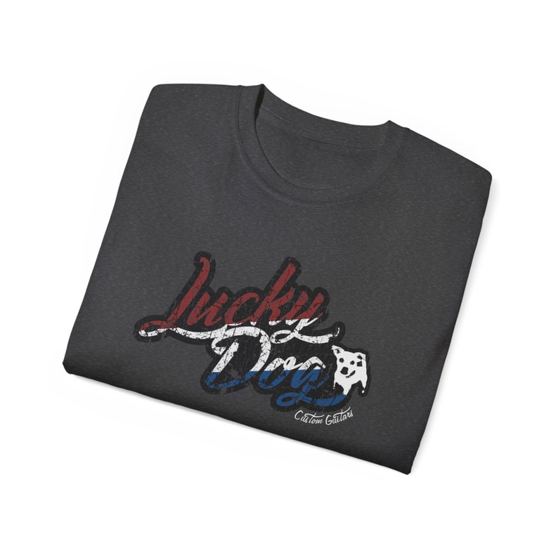 May include: A dark gray t-shirt with a red, white, and blue graphic that says "Lucky Dog" with a small white dog silhouette. The text "Custom Guitars" is below the graphic.