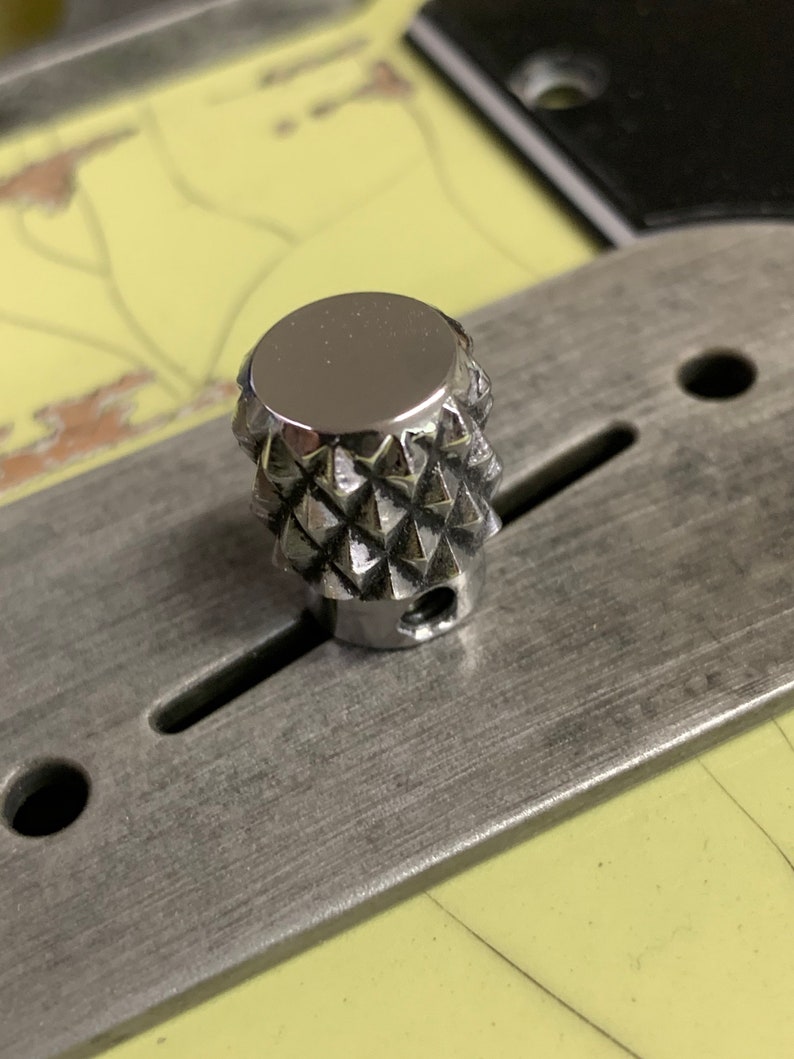 Puede incluir: Un pomo de color plateado en forma de diamante con una parte superior lisa y reflectante. El pomo est&aacute; montado en una superficie met&aacute;lica.