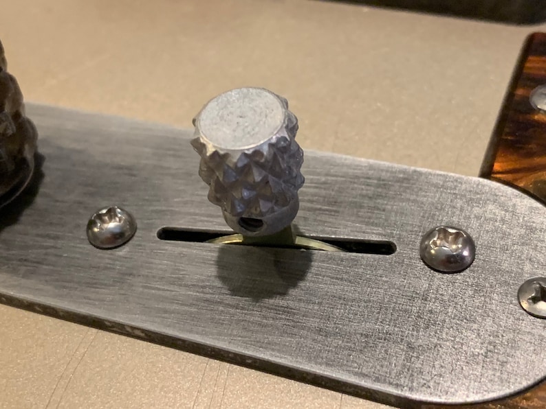Puede incluir: Primer plano de una perilla de guitarra de color plateado con un patr&oacute;n texturizado en forma de diamante. La perilla est&aacute; montada en una placa de metal con dos tornillos.