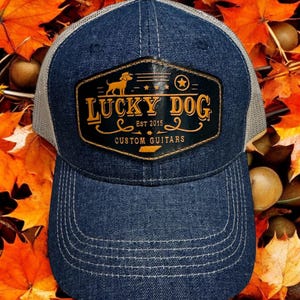 May include: A blue denim trucker hat with a mesh back. The hat has a black leather patch with the text "Lucky Dog" in gold lettering and a small dog silhouette. The patch also includes the text "Est 2015" and "Custom Guitars".