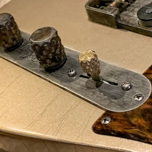Puede incluir: Primer plano de la placa de control de una guitarra con tres botones. Los botones son de metal y tienen un acabado texturizado. La placa es de metal y tiene un acabado cepillado. La guitarra es de color dorado claro.
