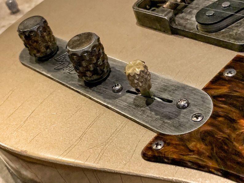 Puede incluir: Primer plano de la placa de control de una guitarra con tres botones. Los botones son de metal y tienen un acabado texturizado. La placa es de metal y tiene un acabado cepillado. La guitarra es de color dorado claro.