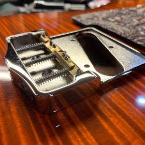 Op de afbeelding: Close-up van een chromen gitaarbrug met drie verstelbare zadels en een messing brugplaat.