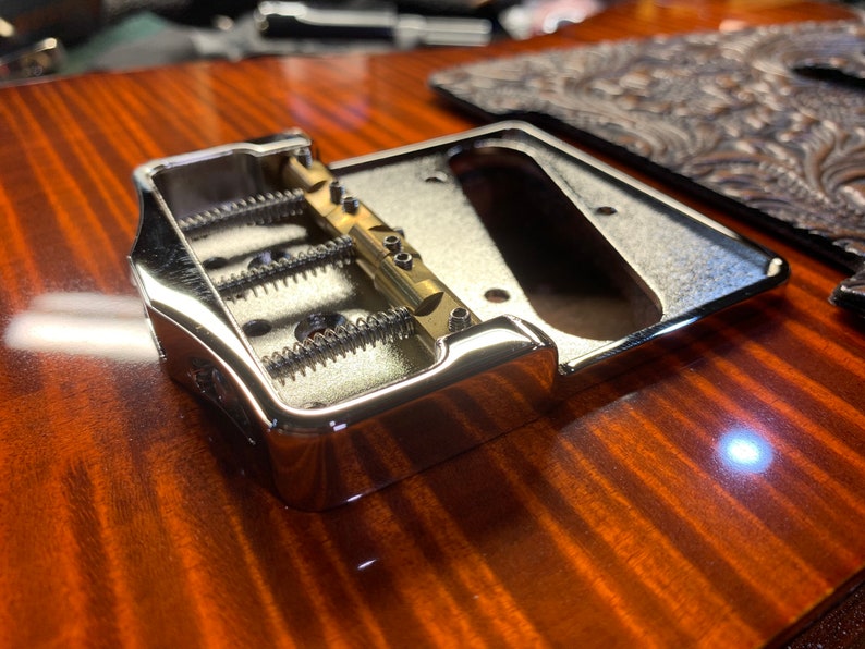 Op de afbeelding: Close-up van een chromen gitaarbrug met drie verstelbare zadels en een messing brugplaat.