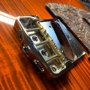 Op de afbeelding: Close-up van een chromen gitaarbrug met een zadel en veren van messing. De brug is gemonteerd op een houten gitaarlichaam.