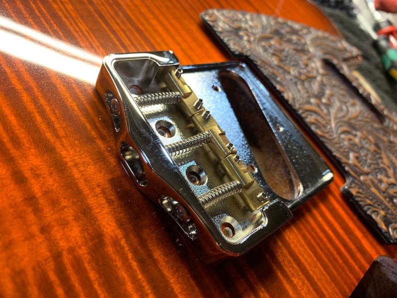 Op de afbeelding: Close-up van een chromen gitaarbrug met een zadel en veren van messing. De brug is gemonteerd op een houten gitaarlichaam.