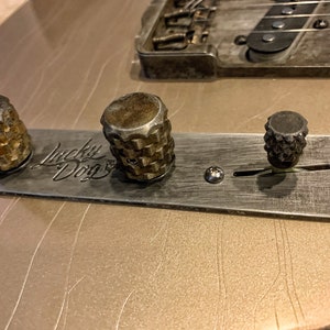 Puede incluir: Primer plano de la placa de control de una guitarra con tres botones y un interruptor de palanca. La placa es de metal y tiene un acabado cepillado. Los botones son de metal y tienen un dise&ntilde;o moleteado. El interruptor de palanca es de metal y tiene una punta de pl&aacute;stico negro. La placa est&aacute; etiquetada como "Lucky Dog".