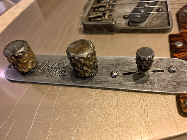 Puede incluir: Primer plano de la placa de control de una guitarra con tres botones y un interruptor de palanca. La placa es de metal y tiene un acabado cepillado. Los botones son de metal y tienen un dise&ntilde;o moleteado. El interruptor de palanca es de metal y tiene una punta de pl&aacute;stico negro. La placa est&aacute; etiquetada como "Lucky Dog".