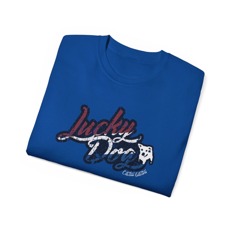 May include: A royal blue t-shirt with a distressed graphic design that says "Lucky Dog" in red, white, and blue with a white silhouette of a dog's head. The text "Custom Guitars" is printed in white below the graphic.