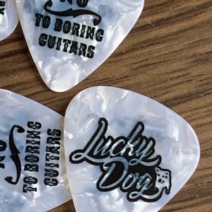 May include: Guitar picks with a pearlescent finish and black text. One pick reads "Just Say No To Boring Guitars." Another says "Lucky Dog" with a dog graphic. The picks are on a wooden surface.
