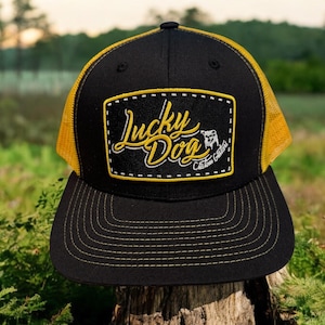 May include: Black and gold trucker hat with a yellow patch that says "Lucky Dog" with a small dog illustration and the text "Get Lucky" underneath.