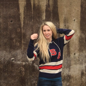 May include: A woman wearing a blue and white striped sweater with red accents and the letters "SA" on the front. She is standing in front of a concrete wall.