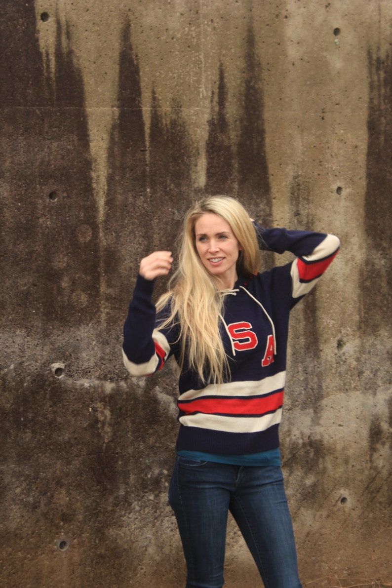 May include: A woman wearing a blue and white striped sweater with red accents and the letters "SA" on the front. She is standing in front of a concrete wall.