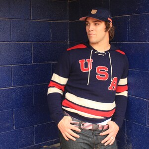 May include: A navy blue and white striped sweater with a red "USA" logo. The sweater has a hood and a drawstring. The person is wearing a blue baseball cap with a white logo.
