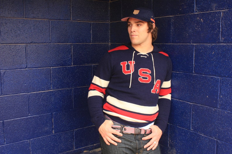 May include: A navy blue and white striped sweater with a red "USA" logo. The sweater has a hood and a drawstring. The person is wearing a blue baseball cap with a white logo.