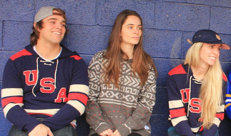 May include: Three people wearing sweaters with the word "USA" on them. The person on the left is wearing a navy blue sweater with white and red stripes on the sleeves. The person in the middle is wearing a gray sweater with a geometric pattern. The person on the right is wearing a navy blue sweater with red and white stripes on the sleeves.
