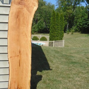 May include: A large, unfinished slab of light brown wood with a natural edge. The wood has visible grain and knots, and is standing upright against a wall. The wood is likely for a table or other furniture project.