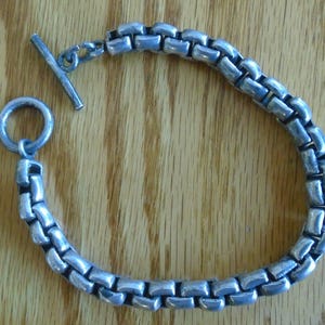 May include: Silver-toned metal bracelet with a rectangular link chain design. The bracelet features a toggle clasp for secure fastening. The jewelry is displayed on a wooden surface, showcasing its metallic sheen and craftsmanship.