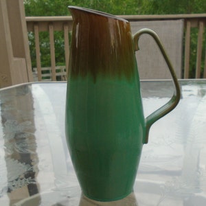 May include: A green and brown ceramic pitcher with a wide mouth and a handle. The pitcher has a glazed finish and a smooth surface.