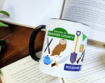 May include: White ceramic mug with a black handle. The mug features a colorful illustration of a welcome sign to Midsomer County, a brown owl, a teapot, a knife, a shovel, and a jar with a skull and crossbones. The text "Welcome to Midsomer County" is printed on the sign.