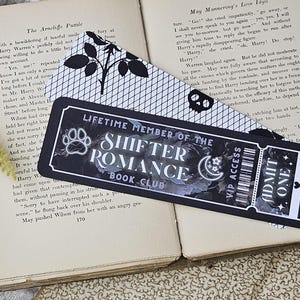 May include: Two bookmarks rest on an open book. One is black and white with a floral design. The other is a black rectangular bookmark with the text "LIFETIME MEMBER OF THE SHIFTER ROMANCE BOOK CLUB". It also has a paw print, moon, stars, and "ADMIT ONE" text.