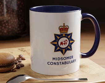 May include: White ceramic mug with a blue rim and handle. The mug features a gold star with a red badger inside, a gold crown above the star, and the text "Midsomer Constabulary".
