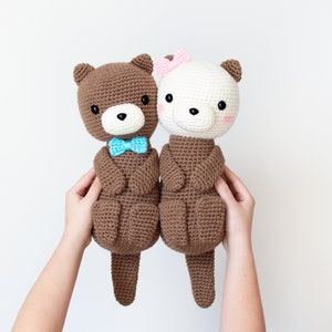 May include: Two crocheted otter plushies, one brown with a blue bow tie and one white with a pink bow. The otters are holding hands and have black eyes and noses.