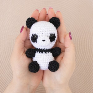 Op de afbeelding: Een klein gehaakt pandabeer speelgoed met zwarte oren, ogen en poten, en een wit lichaam. De panda wordt in de handpalmen van twee handen gehouden.