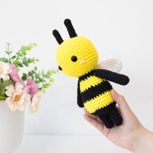 May include: A crocheted bee plush toy with black and yellow stripes, white wings, and black antennae. The bee has black eyes and a pink blush on its cheeks.