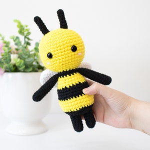 May include: A crocheted bee plush toy with black and yellow stripes, black antennae, and white wings. The bee has black eyes and a pink blush.