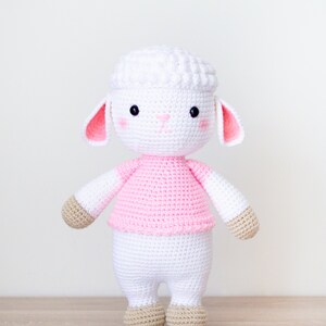 May include: A white crocheted lamb wearing a pink shirt. The lamb has a fluffy white head with black eyes and a pink nose. The lamb is standing on a wooden surface.