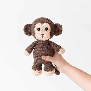 May include: A brown crocheted monkey toy with black eyes and a pink nose. The monkey is being held by a hand.