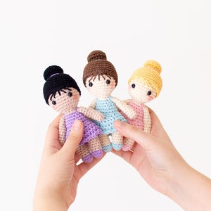 May include: Three crocheted dolls dressed in ballerina outfits. The dolls are wearing pink, blue, and purple dresses. They have brown, black, and blonde hair.