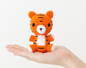 May include: A small orange and black crocheted tiger soft toy with white accents. The tiger is sitting on a hand.