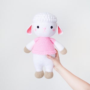 May include: A white crocheted lamb wearing a pink shirt. The lamb has brown hooves and black eyes.