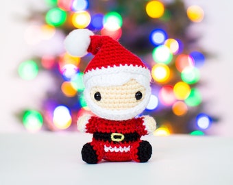 Puede incluir: Un muñeco de Papá Noel de crochet con un gorro rojo, barba blanca y botas negras. El muñeco está sentado sobre una superficie blanca con un fondo borroso de luces navideñas.