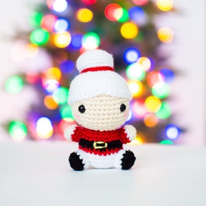 May include: A crocheted Santa Claus doll with a white hat and red trim. The doll has a beige face, black eyes, and a red and black outfit with a gold buckle. The background is a blurred display of colorful Christmas lights.