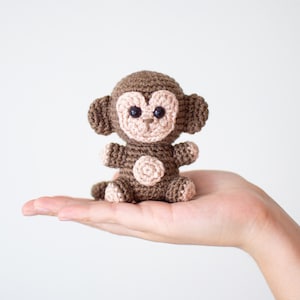 May include: A small brown crocheted monkey toy with pink accents on the face and belly. The monkey is sitting in the palm of a hand.