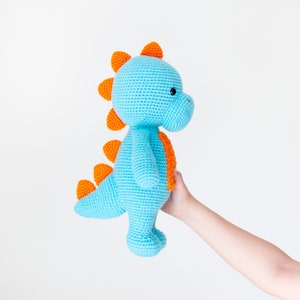 Pode incluir: Um brinquedo de dinossauro azul de crochê com espinhos laranja e olhos pretos. O dinossauro está sendo segurado por uma mão.