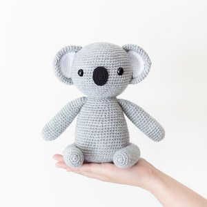 May include: A gray crocheted koala plush toy with black eyes and a black nose. The koala is sitting on a hand.