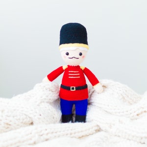 May include: A crocheted toy soldier with a red and black uniform, a black hat, and a white face. The toy is sitting on a white knitted blanket.