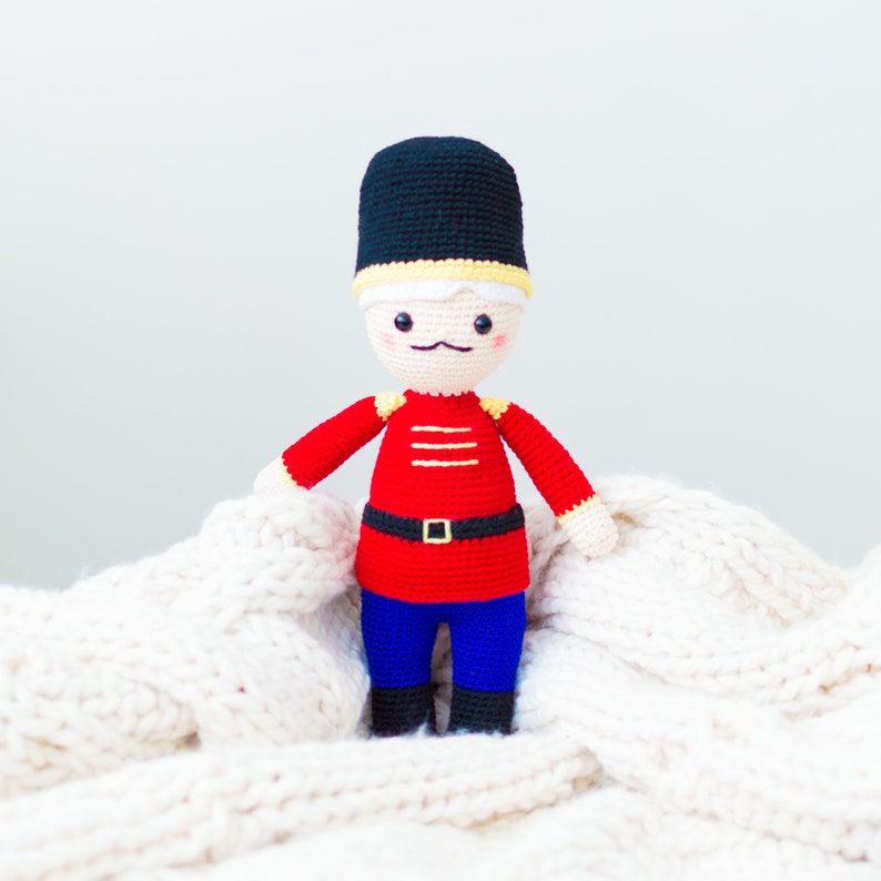 May include: A crocheted toy soldier with a red and black uniform, a black hat, and a white face. The toy is sitting on a white knitted blanket.