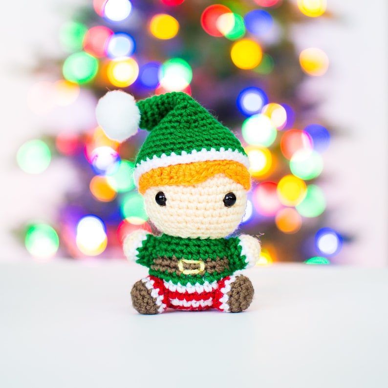 May include: A crocheted elf doll with a green hat, white trim, and orange hair. The doll is wearing a green shirt, brown belt, and red and white striped pants. The doll is sitting on a white surface with a blurry background of Christmas lights.